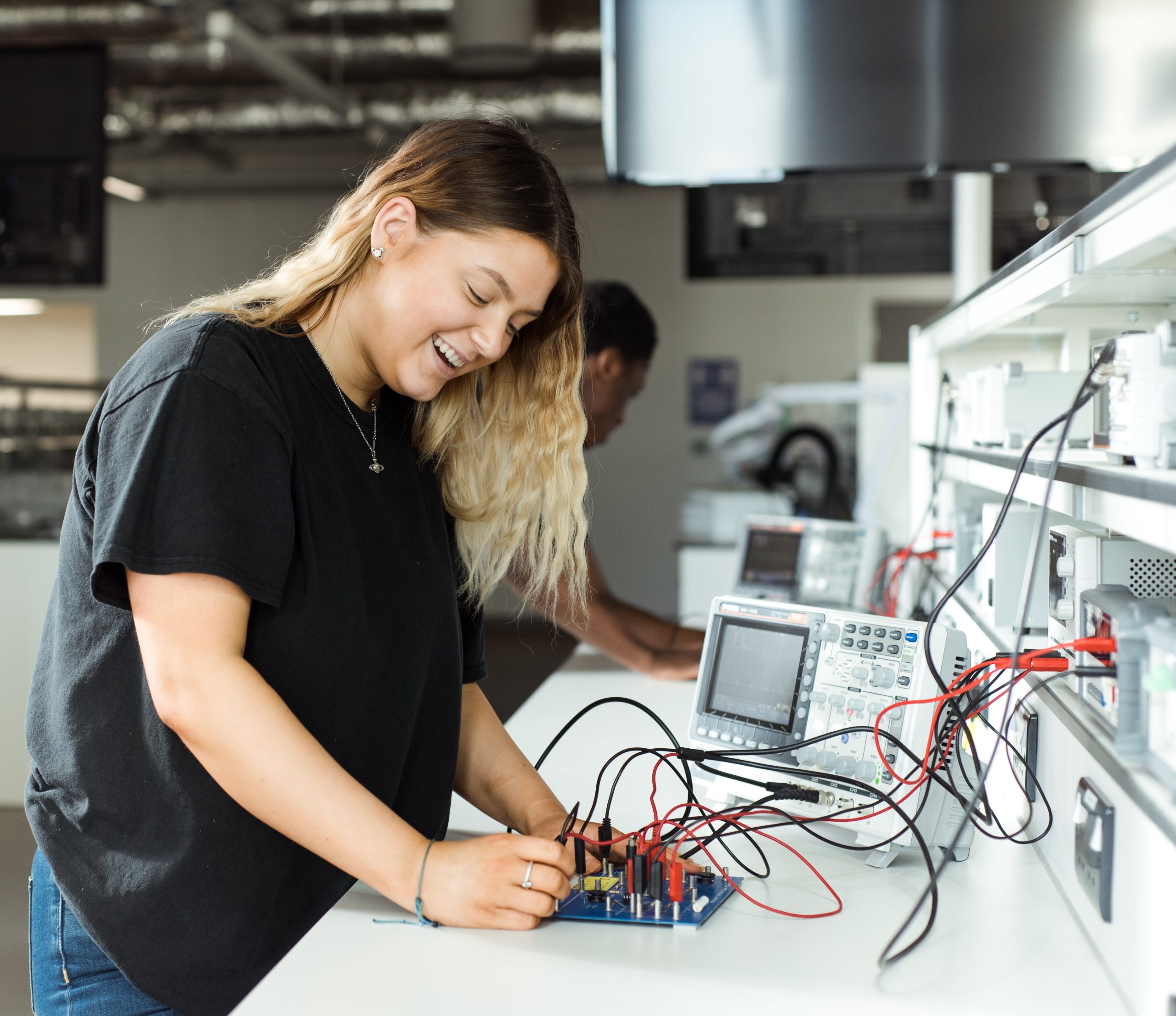 Student happily working on a circuit board with another student in the back.